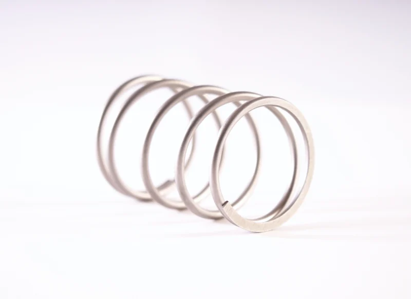 Primer plano de un muelle de compresión de acero inoxidable con espiras de gran diámetro y acabado mate, orientado diagonalmente sobre un fondo blanco neutro.