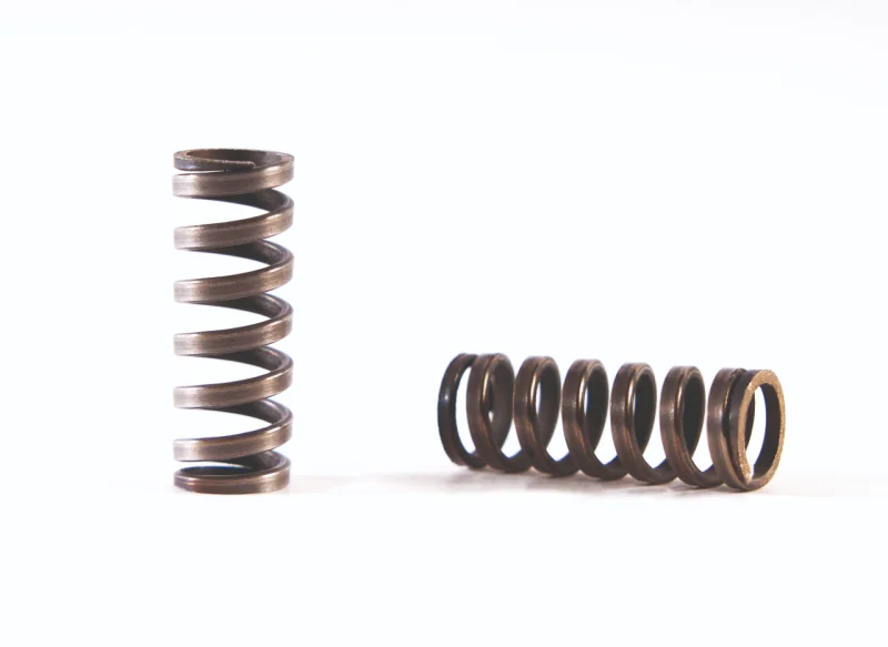 Dos muelles de compresión de acero oscuro con acabado pavonado sobre fondo blanco: uno colocado verticalmente y otro tumbado de forma horizontal, mostrando su robustez.