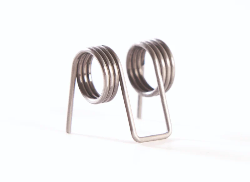 Muelle de torsión doble de acero sobre fondo blanco, con dos bobinas helicoidales conectadas por un puente central en forma de U y brazos de apoyo laterales.