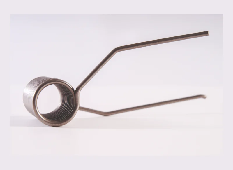 Muelle de torsión de acero inoxidable sobre fondo blanco, compuesto por un cuerpo helicoidal central y dos brazos metálicos rectos de diferentes longitudes extendidos lateralmente.