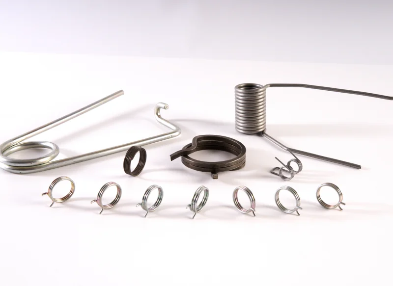 Primer plano de un muelle de torsión de hilo plano sobre fondo blanco, mostrando sus espiras rectangulares de acero oscuro y dos patas cortas de apoyo.